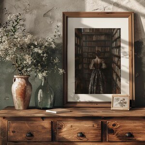 May include: A framed print of a woman standing in a library, looking at bookshelves filled with books. The woman is wearing a long, dark dress. The print is on a wooden table with a vase of white flowers and a small framed print of a plant.