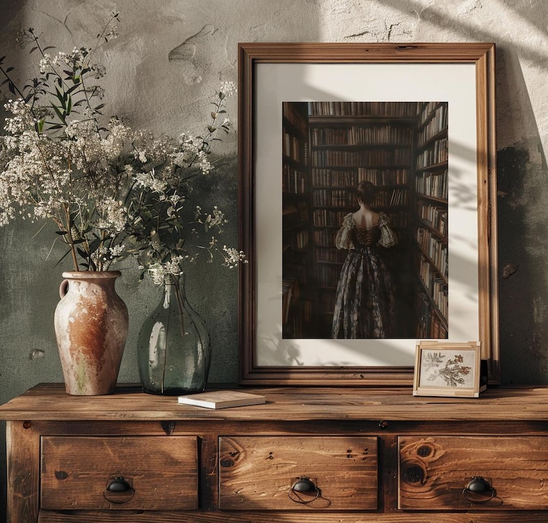 May include: A framed print of a woman standing in a library, looking at bookshelves filled with books. The woman is wearing a long, dark dress. The print is on a wooden table with a vase of white flowers and a small framed print of a plant.