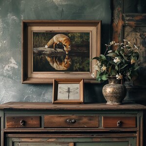 May include: A framed painting of a fox drinking from a pond, with a smaller framed dragonfly print below. The artwork is displayed on a wooden cabinet with a decorative vase of flowers. The color palette includes browns, greens, and gold.