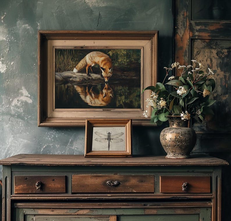 May include: A framed painting of a fox drinking from a pond, with a smaller framed dragonfly print below. The artwork is displayed on a wooden cabinet with a decorative vase of flowers. The color palette includes browns, greens, and gold.