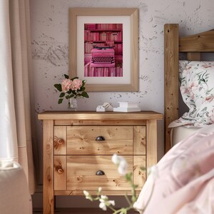 K&ouml;nnte beinhalten: Eine rosafarbene Vintage-Schreibmaschine steht auf einem Regal mit rosafarbenen B&uuml;chern in einem gerahmten Bild &uuml;ber einem Holz-Nachttisch mit zwei Schubladen. Der Nachttisch steht neben einem Bett mit einem rosafarbenen und wei&szlig;en Kissen mit Blumenmuster und einer rosafarbenen Decke.