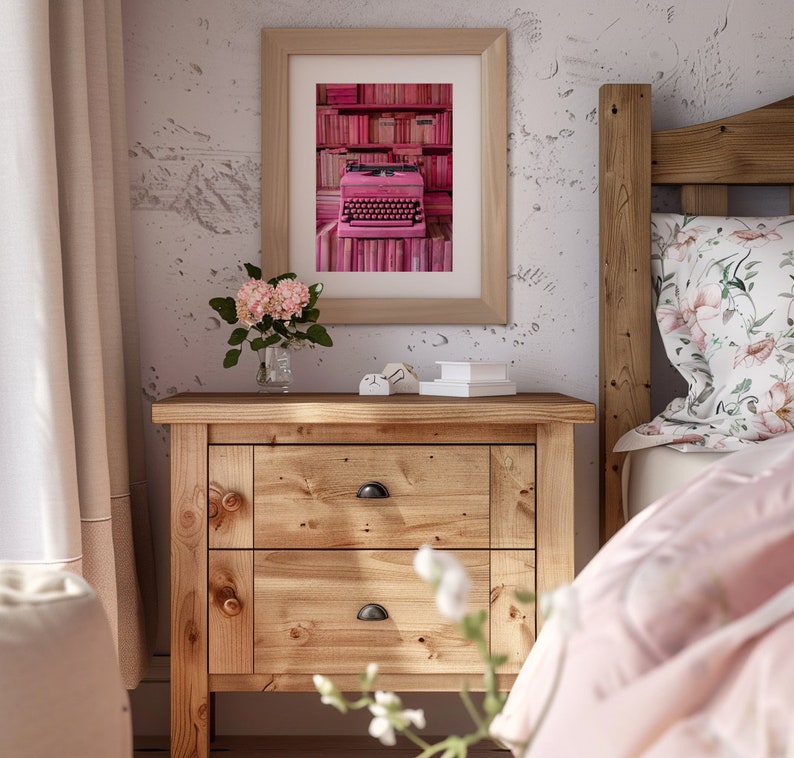 K&ouml;nnte beinhalten: Eine rosafarbene Vintage-Schreibmaschine steht auf einem Regal mit rosafarbenen B&uuml;chern in einem gerahmten Bild &uuml;ber einem Holz-Nachttisch mit zwei Schubladen. Der Nachttisch steht neben einem Bett mit einem rosafarbenen und wei&szlig;en Kissen mit Blumenmuster und einer rosafarbenen Decke.