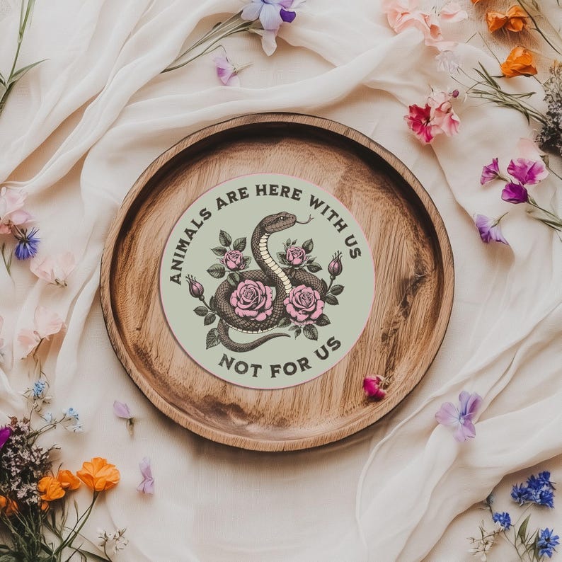 May include: A round sticker with the text "ANIMALS ARE HERE WITH US NOT FOR US" encircling a central design. The design features a snake intertwined with pink roses and green leaves. The sticker is on a wooden tray.