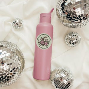 May include: A pink water bottle with a flip-top lid and a circular sticker that reads "ANIMALS ARE HERE WITH US NOT FOR US" with a snake and floral design. The bottle is surrounded by several disco balls.