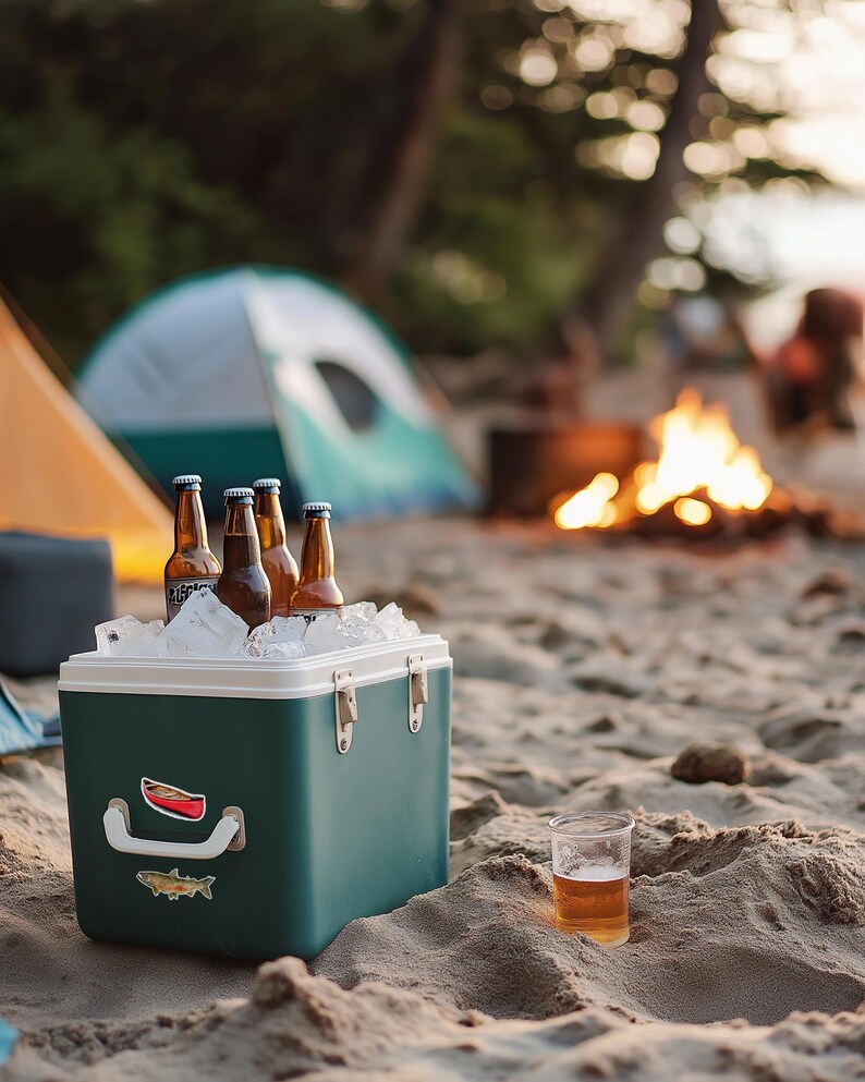 May include: A dark green rectangular cooler filled with ice and four brown glass beer bottles. The cooler features a white handle, two metal latches, and two stickers depicting a red canoe and a fish. A clear plastic cup with beer sits nearby.