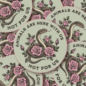 May include: Round sticker with a snake and pink roses design. The text "ANIMALS ARE HERE WITH US" and "NOT FOR US" is printed around the edge of the sticker. The snake is brown and the roses are pink.