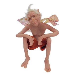 May include: A small, handmade fairy figurine with white hair, a wrinkled face, and a mischievous smile. The fairy is wearing orange shorts and has delicate wings.