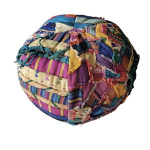 May include: A round, patchwork pouf in a variety of colors including blue, purple, yellow, and green. The pouf is made of stitched fabric pieces, creating a textured surface. It has a rope-like trim around the top.