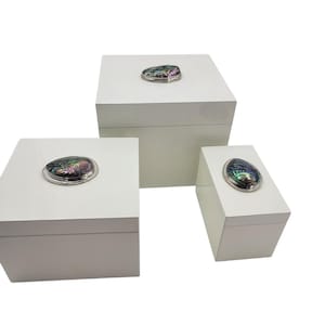 May include: Three white square boxes with abalone shell accents. The boxes are stacked on top of each other, with the largest box on the bottom and the smallest box on top.