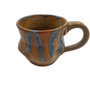 Puede incluir: Taza de cerámica hecha a mano con un diseño texturizado único. La taza presenta una base marrón con goteos de esmalte azul y naranja. El asa tiene un diseño simple y curvado, y la taza tiene una forma orgánica e irregular.