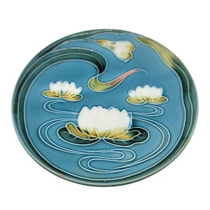 Może przedstawiać: Okrągły ceramiczny talerz z turkusowym tłem przedstawiającym wzór lilii wodnych. Talerz ma białe lilie wodne z żółtymi środkami, zielone liście lilii i białe faliste linie. Obręcz jest ciemniejszego turkusu.