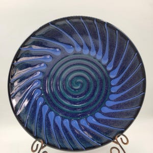 May include: A handcrafted ceramic plate with a spiral design. The plate features a dark blue background with a lighter blue spiral in the center. Light blue teardrop shapes radiate from the spiral. The plate is displayed on a decorative metal stand.