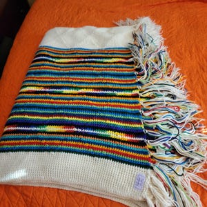 May include: A handmade, colorful crochet blanket with horizontal stripes in various colors, including red, blue, yellow, and green. The blanket has a white top and bottom border, and a fringe of colorful yarn. The blanket is on an orange surface.