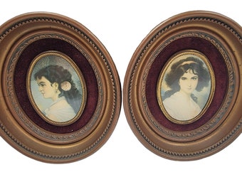 May include: Two antique oval portrait frames. Each frame has a gold-colored, ornate outer frame, a burgundy velvet inner frame, and a portrait of a woman. The portraits are in oval frames with a gold border.