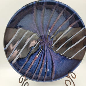 May include: A handmade ceramic plate with a spiral design. The plate features a dark blue rim and a center that swirls with shades of purple, brown, and white. The plate is displayed on a decorative metal stand.