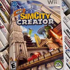 May include: A Wii game case for "SimCity Creator" featuring a cityscape with a red fire truck, a green dragon-like creature, and the EA logo. The game case is surrounded by other Wii game cases.