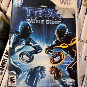 May include: A Wii game case for "Tron: Evolution - Battle Grids" featuring two figures in black and blue suits with glowing accents. The case has the Disney logo and is rated "Everyone 10+". The game is compatible with Wii MotionPlus.