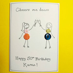 May include: A white card with black ink that says "Cheers me dears" and "Happy 50th Birthday Karen!"  Two stick figures are drawn on the card, each holding a glass of champagne.  The stick figures are decorated with colorful buttons.