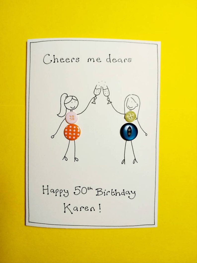 May include: A white card with black ink that says "Cheers me dears" and "Happy 50th Birthday Karen!"  Two stick figures are drawn on the card, each holding a glass of champagne.  The stick figures are decorated with colorful buttons.