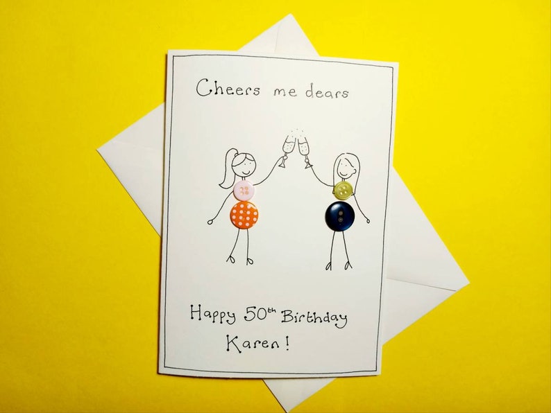 May include: A white card with a hand-drawn illustration of two people toasting with champagne glasses. The card says "Cheers me dears" and "Happy 50th Birthday Karen!" with colorful buttons decorating the illustration.