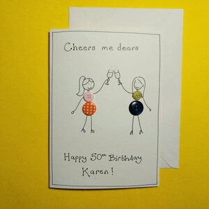 May include: A white card with a hand-drawn illustration of two people toasting with champagne glasses. The card says "Cheers me dears" and "Happy 50th Birthday Karen!" with colorful buttons attached to the illustration.