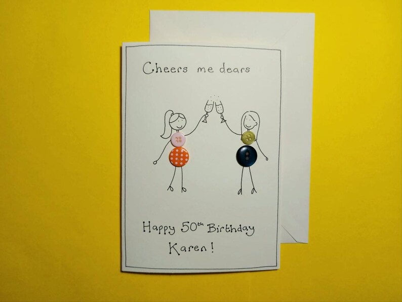 May include: A white card with a hand-drawn illustration of two people toasting with champagne glasses. The card says "Cheers me dears" and "Happy 50th Birthday Karen!" with colorful buttons attached to the illustration.