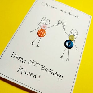 May include: A handmade birthday card with a hand-drawn illustration of two people toasting with glasses of champagne. The card is decorated with colorful buttons and the text "Cheers me dears Happy 50th Birthday Karen!"