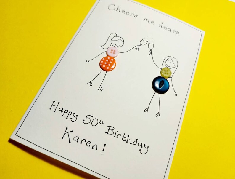 May include: A handmade birthday card with a hand-drawn illustration of two people toasting with glasses of champagne. The card is decorated with colorful buttons and the text "Cheers me dears Happy 50th Birthday Karen!"