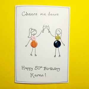May include: A white card with a hand-drawn illustration of two people toasting with champagne glasses. The card says "Cheers me dears" and "Happy 50th Birthday Karen!" The illustration is decorated with colorful buttons.