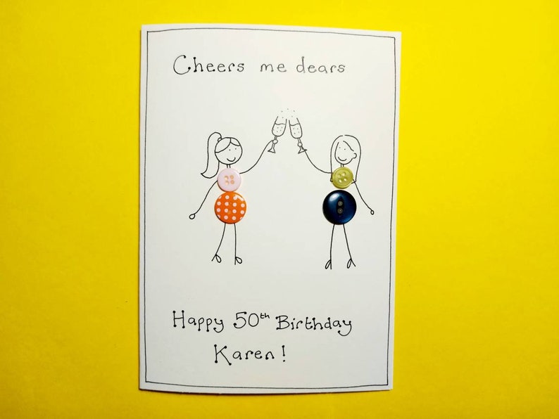 May include: A white card with a hand-drawn illustration of two people toasting with champagne glasses. The card says "Cheers me dears" and "Happy 50th Birthday Karen!" The illustration is decorated with colorful buttons.
