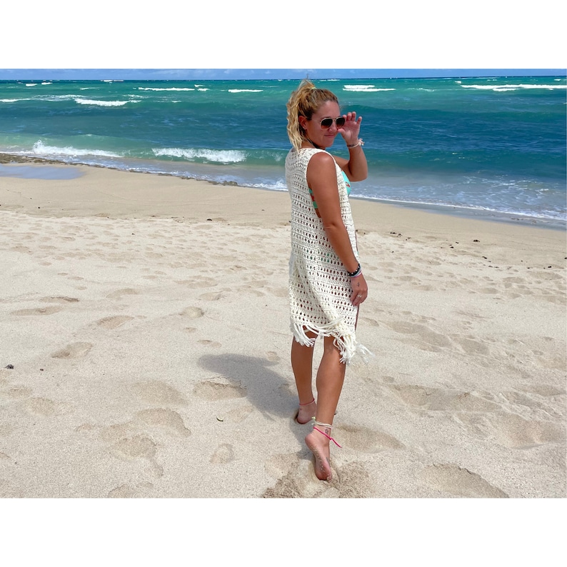 May include: A woman wearing a white crochet cover-up with fringe on a sandy beach. The ocean is in the background.