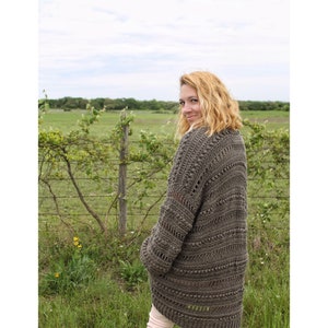May include: A woman wearing a brown crocheted cardigan with a textured design. The cardigan has a loose fit and a long length. The woman is standing in a field with green grass and a fence in the background.