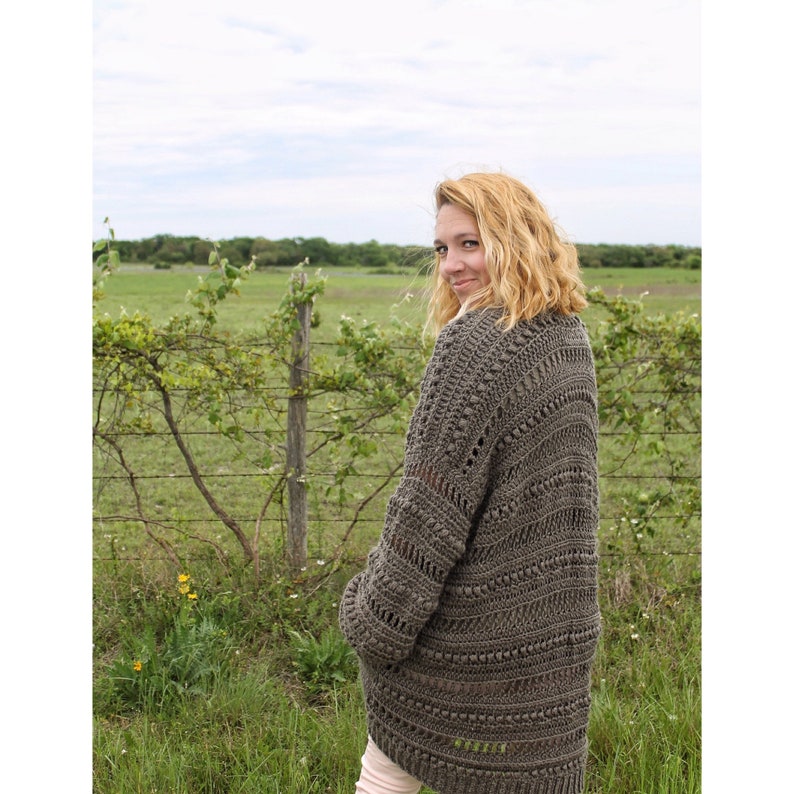 May include: A woman wearing a brown crocheted cardigan with a textured design. The cardigan has a loose fit and a long length. The woman is standing in a field with green grass and a fence in the background.