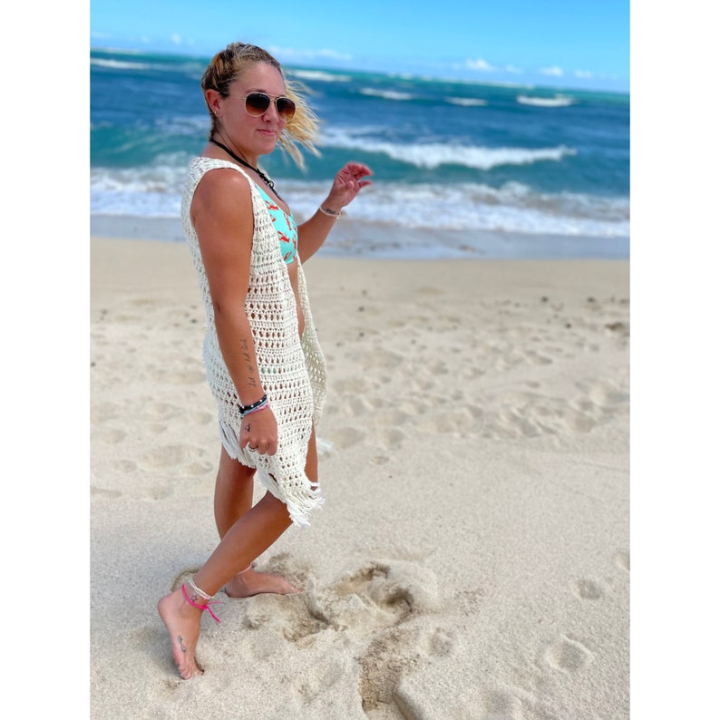 May include: A woman wearing a white crochet cover-up over a turquoise bikini top walks on a sandy beach. The ocean is in the background.