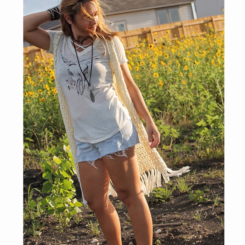 May include: A woman wearing a white crochet vest with fringe, a white t-shirt with a skull design, and denim shorts. She is standing in a field of yellow flowers.