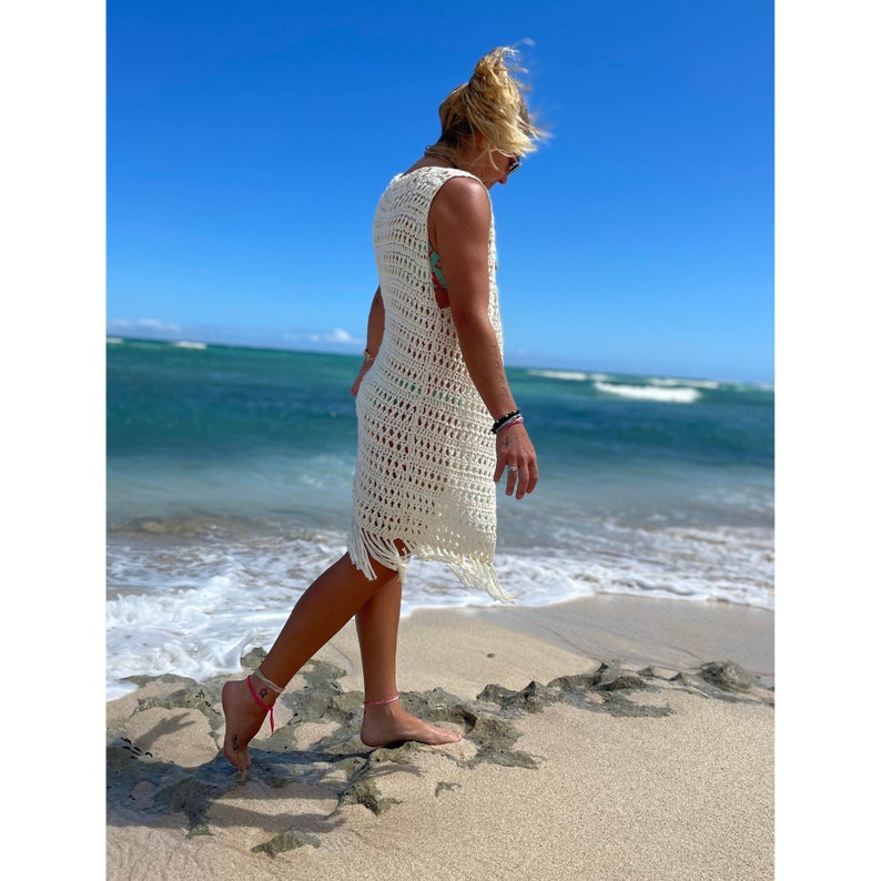 May include: A woman wearing a white crochet cover-up with fringe details walks along a sandy beach with ocean waves in the background.