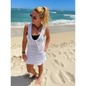 K&ouml;nnte beinhalten: Ein wei&szlig;es H&auml;kel-Strandkleid mit V-Ausschnitt und lockerer Passform. Das Kleid wird von einer Person getragen, die auf einem Sandstrand mit dem Meer im Hintergrund steht.