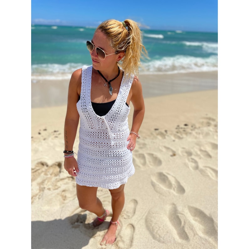 K&ouml;nnte beinhalten: Ein wei&szlig;es H&auml;kel-Strandkleid mit V-Ausschnitt und lockerer Passform. Das Kleid wird von einer Person getragen, die auf einem Sandstrand mit dem Meer im Hintergrund steht.