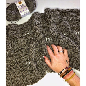 May include: A close-up of a brown crocheted blanket with a white yarn pattern. The blanket is made with a basic stitch and is lying on a white surface. A hand with a ring and a tattoo is resting on the blanket.