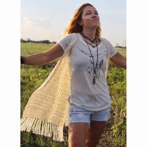 May include: A woman wearing a white crochet vest with fringe, a white t-shirt with a graphic print, and denim shorts. She is standing in a field of grass.