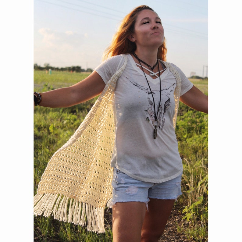 May include: A woman wearing a white crochet vest with fringe, a white t-shirt with a graphic print, and denim shorts. She is standing in a field of grass.
