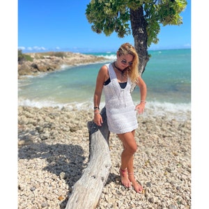 K&ouml;nnte beinhalten: Eine Frau in einem wei&szlig;en H&auml;kelkleid und Sonnenbrille steht auf einem Treibholzstamm an einem Strand. Der Strand ist felsig und das Wasser ist t&uuml;rkisfarben.