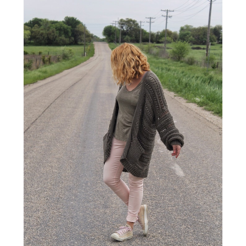 May include: A woman wearing a long, dark green cardigan with a loose, open knit pattern. She is wearing pink pants and white sneakers. She is standing on a paved road with green grass on either side.
