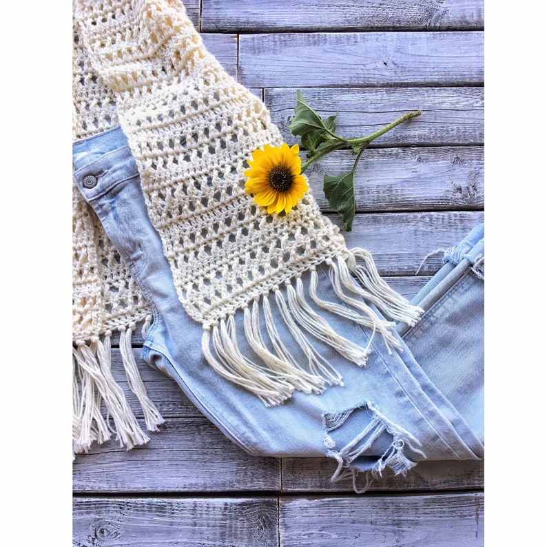 May include: A white crocheted scarf with fringe, a pair of light blue jeans, and a yellow sunflower on a white wooden background.