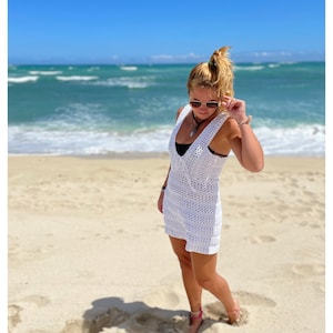K&ouml;nnte beinhalten: Eine Frau in einem wei&szlig;en H&auml;kelkleid am Strand. Sie steht auf dem Sand mit dem Meer im Hintergrund.