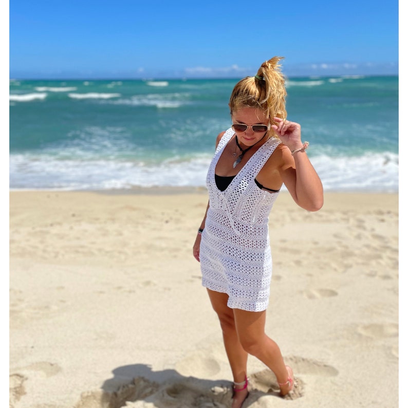 K&ouml;nnte beinhalten: Eine Frau in einem wei&szlig;en H&auml;kelkleid am Strand. Sie steht auf dem Sand mit dem Meer im Hintergrund.