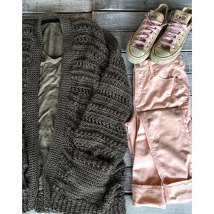 May include: A brown crocheted cardigan with a light brown t-shirt underneath. Pink jeans and a pair of tan canvas sneakers with pink laces are also pictured.