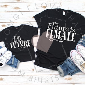 Peut inclure: Deux t-shirts noirs avec du texte blanc. Le plus grand t-shirt dit "The Future is Female". Le plus petit t-shirt dit "I'm The Future".