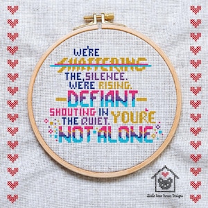 May include: Cross-stitch embroidery in a wooden hoop with the words "We're Shattering the Silence. We're Rising. Defiant. Shouting in the Quiet. You're Not Alone." The design features colorful text and a border of red hearts.
