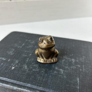 May include: A small, brass frog figurine with dark eyes. The frog is sitting upright, with its front legs close to its body. The figurine has a vintage look and is placed on a dark, textured surface.